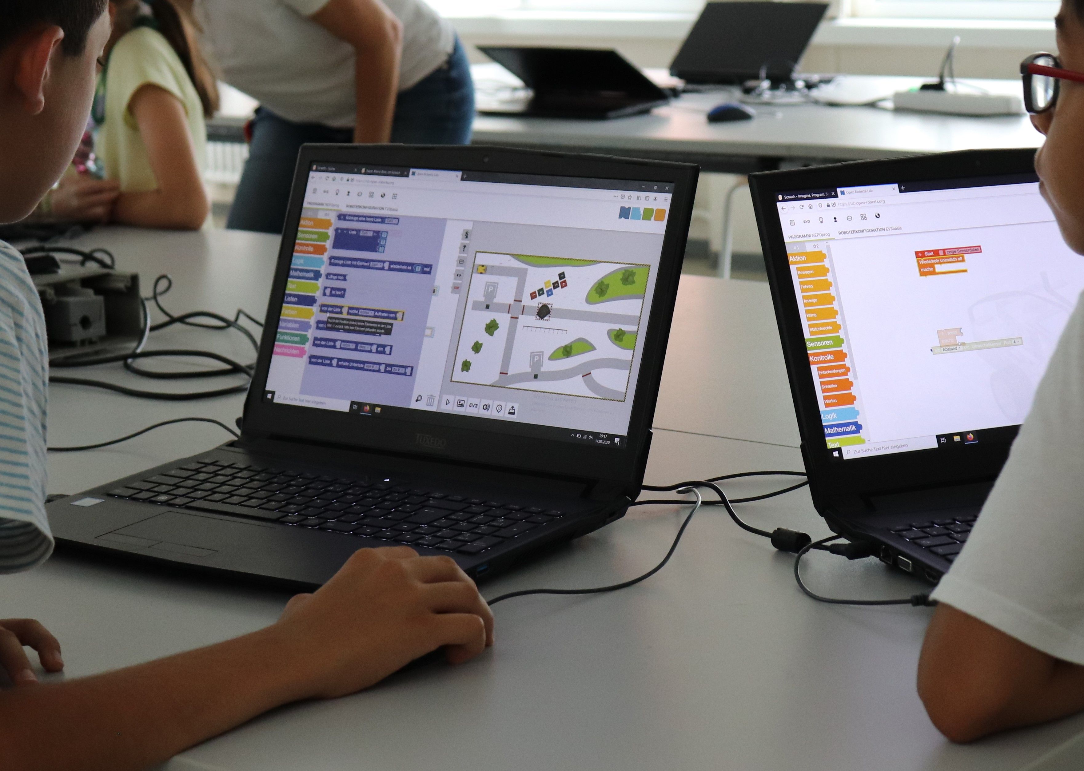 Zwei Jungen programmieren an ihren Laptops mit farbigen Blöcken, auf dem Bildschirm fährt ein simulierter Roboter über eine Karte