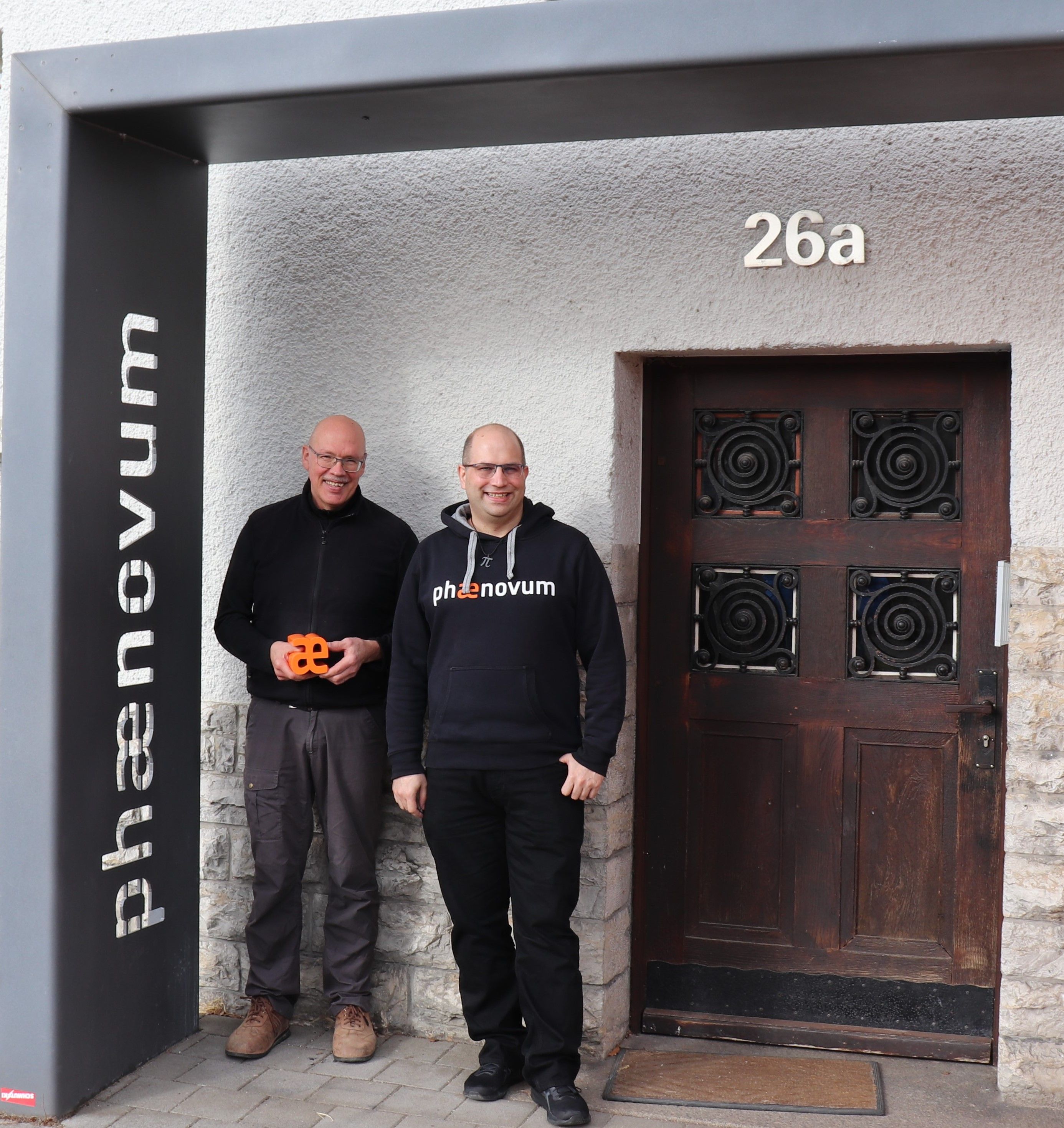 Zwei Männer stehen vor dem Eingang des phænovum in der Hausnummer 26a, einer von ihnen hält das orange æ in den Händen