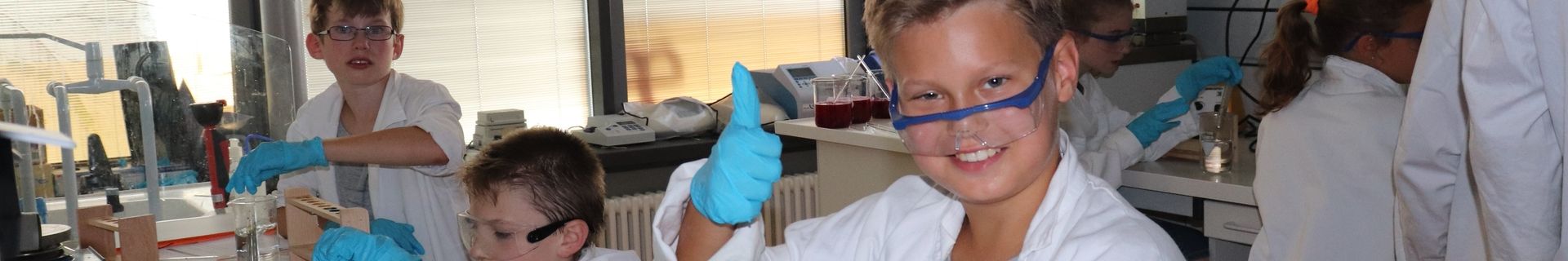 Kinder in Laborkitteln, Schutzbrillen und blauen Handschuhen experimentieren an Labortischen, einer von ihnen hebt den Daumen