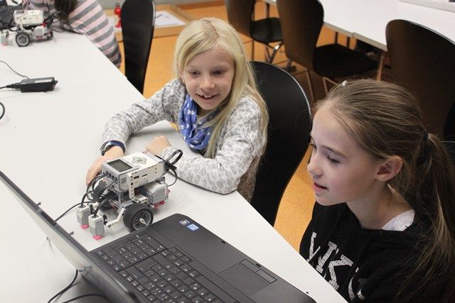 Zwei Mädchen sitzen am Tisch vor einem Lego-Mindstorms-Roboter und einem Laptop und beobachten ihn