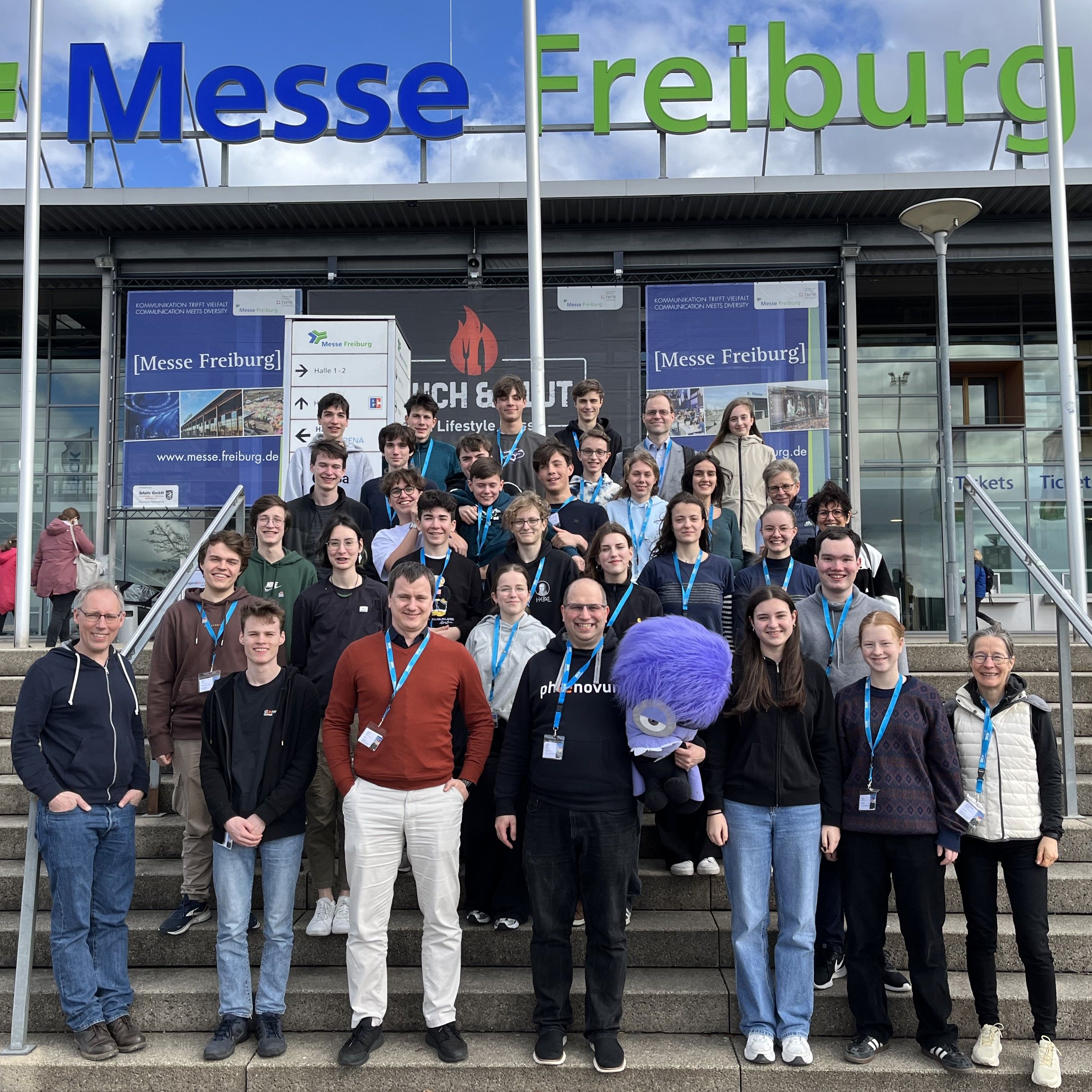 Große Gruppe Jugendlicher und Betreuender auf der Treppe vor dem Eingang der Messe Freiburg, in der Mitte ein lila Plüschmonster