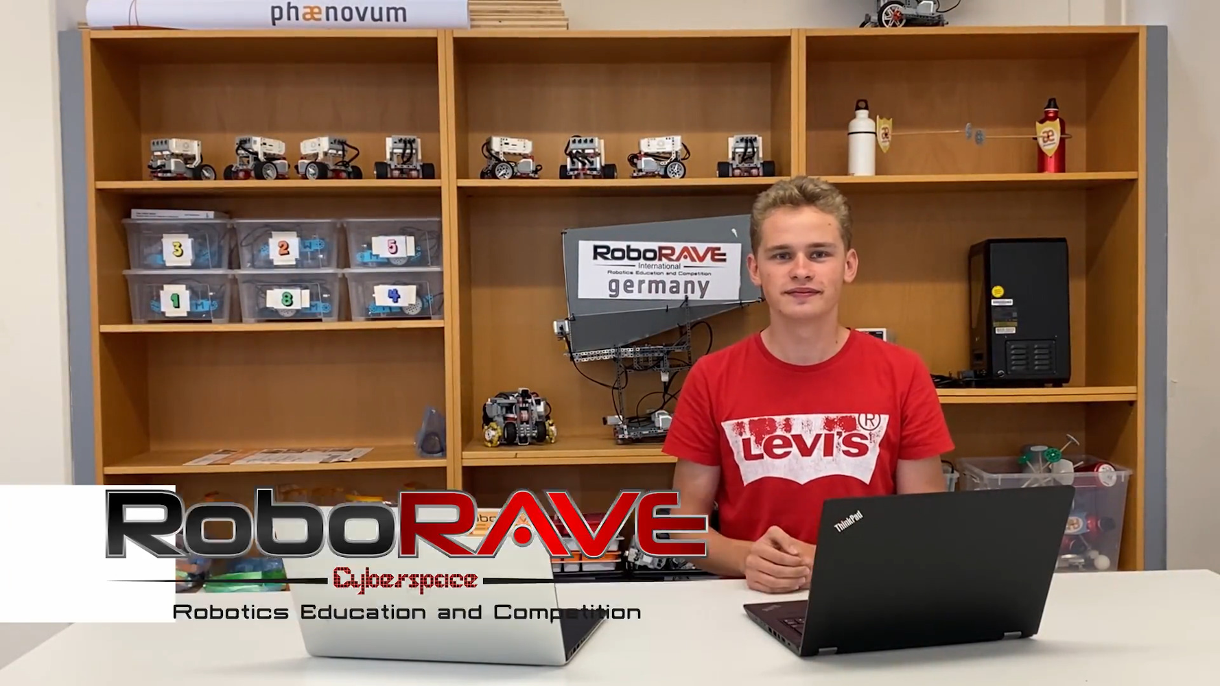 Standbild aus dem Erklärvideo: ein Jugendlicher sitzt mit zwei Laptops vor den Regalen voller Roboter, eingeblendet das Logo des RoboRAVE Cyberspace
