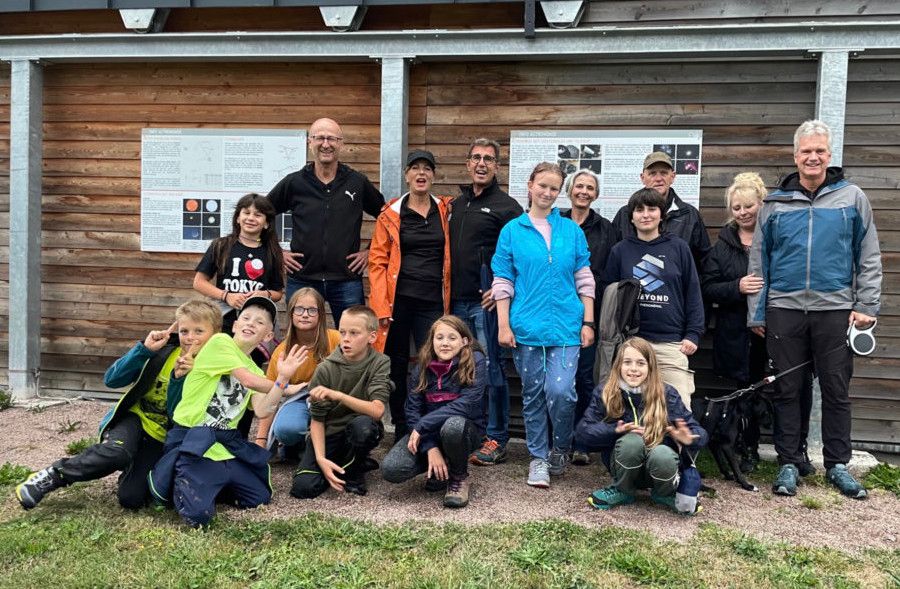 Kinder und Erwachsene stehen und hocken für ein Gruppenfoto vor einer Holzhütte mit zwei Schautafeln zur Astronomie