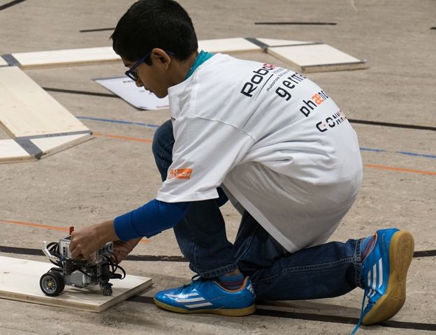 Ein Junge im Wettbewerbs-T-Shirt kniet auf dem Hallenboden und setzt seinen Roboter auf das Startbrett