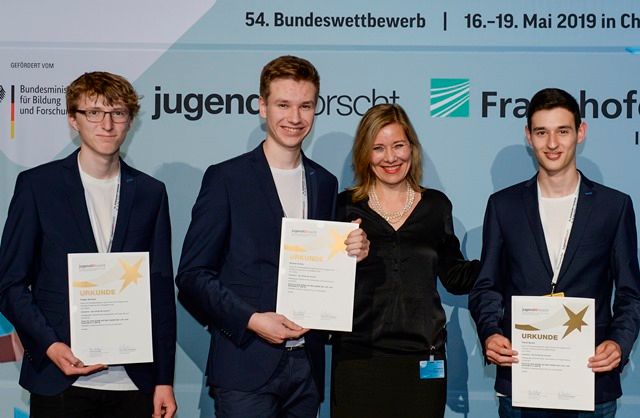 Drei Preisträger halten ihre Urkunden vor der Wand des 54. Bundeswettbewerbs von Jugend forscht im Mai 2019, neben ihnen eine Erwachsene