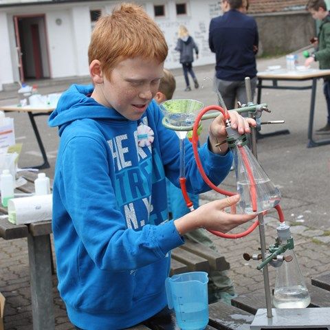 Ein Junge im blauen Kapuzenpullover filtriert auf dem Schulhof eine Flüssigkeit durch einen Trichter in einen Erlenmeyerkolben