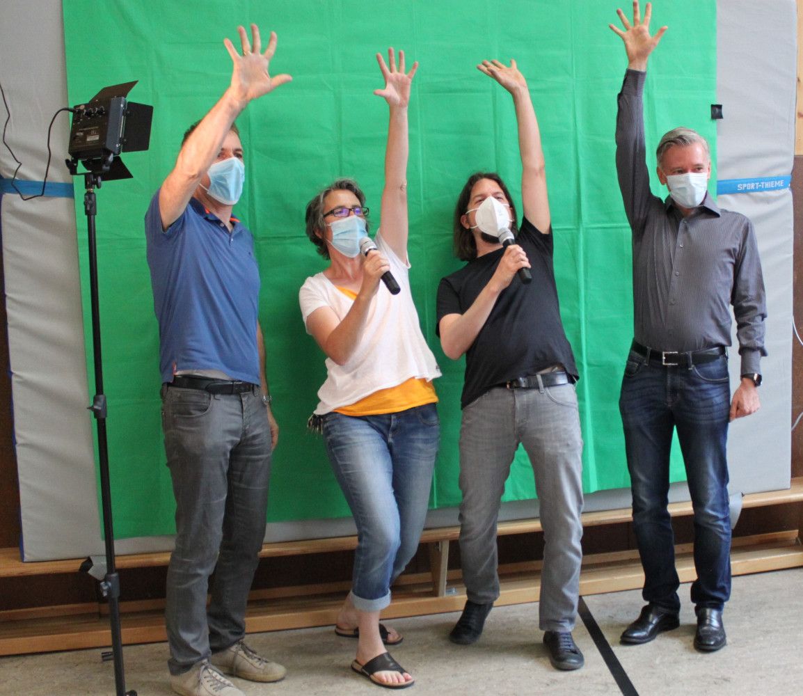 Vier Erwachsene mit Mund-Nasen-Schutz heben vor einer grünen Greenscreen-Wand die Hand, zwei von ihnen halten Mikrofone