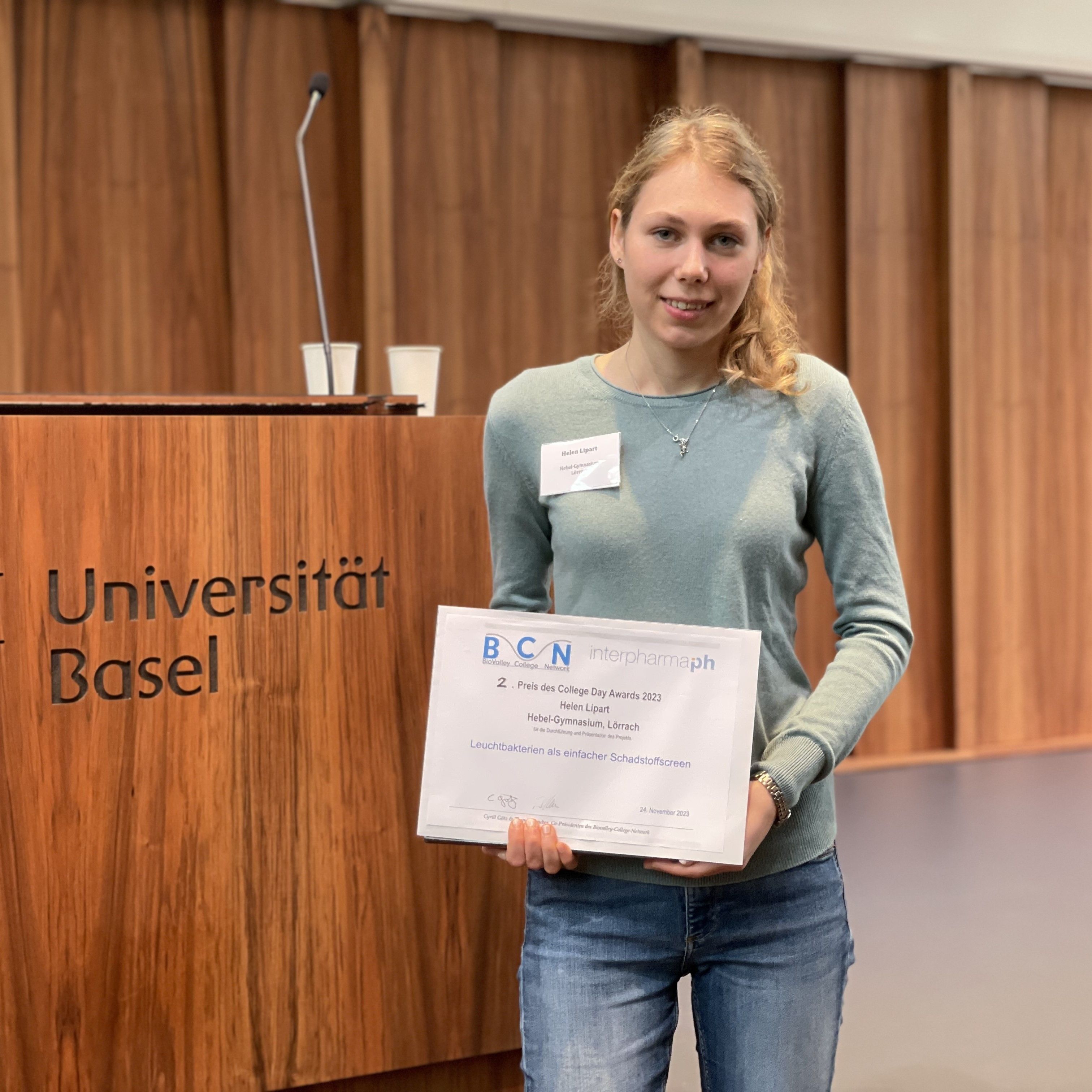 Eine junge Frau hält die Urkunde für den zweiten Preis des College Day Awards 2023 vor dem Rednerpult der Universität Basel