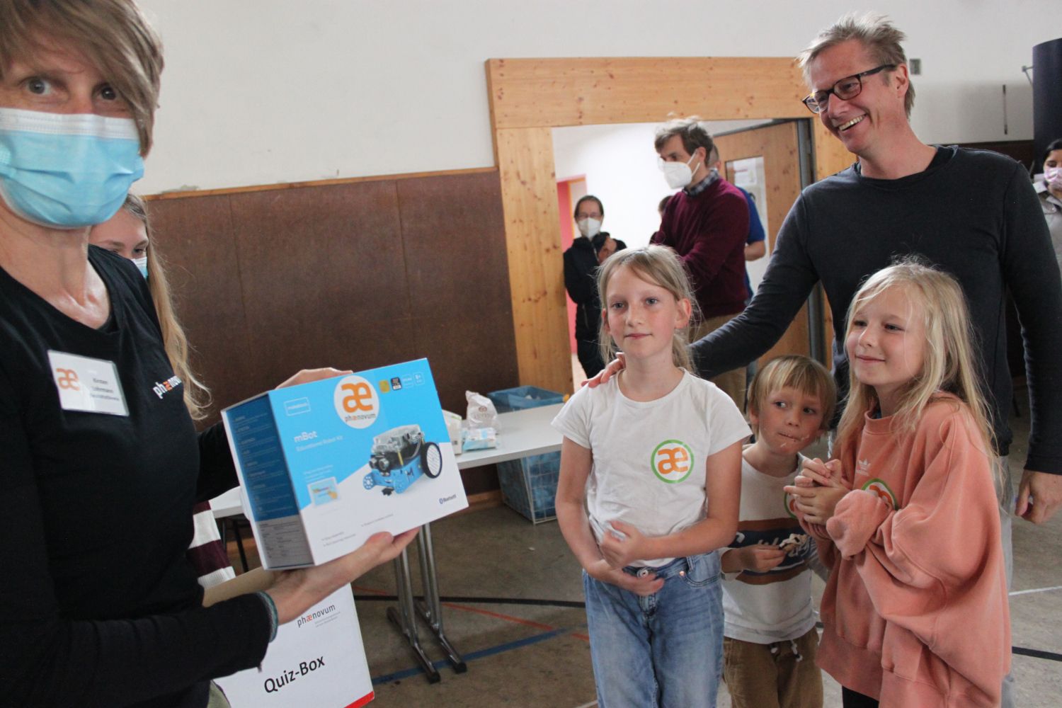 Eine Frau hält einen Roboter-Bausatz und eine Quiz-Box in die Kamera, daneben stehen drei Kinder und ein Mann in einer Halle