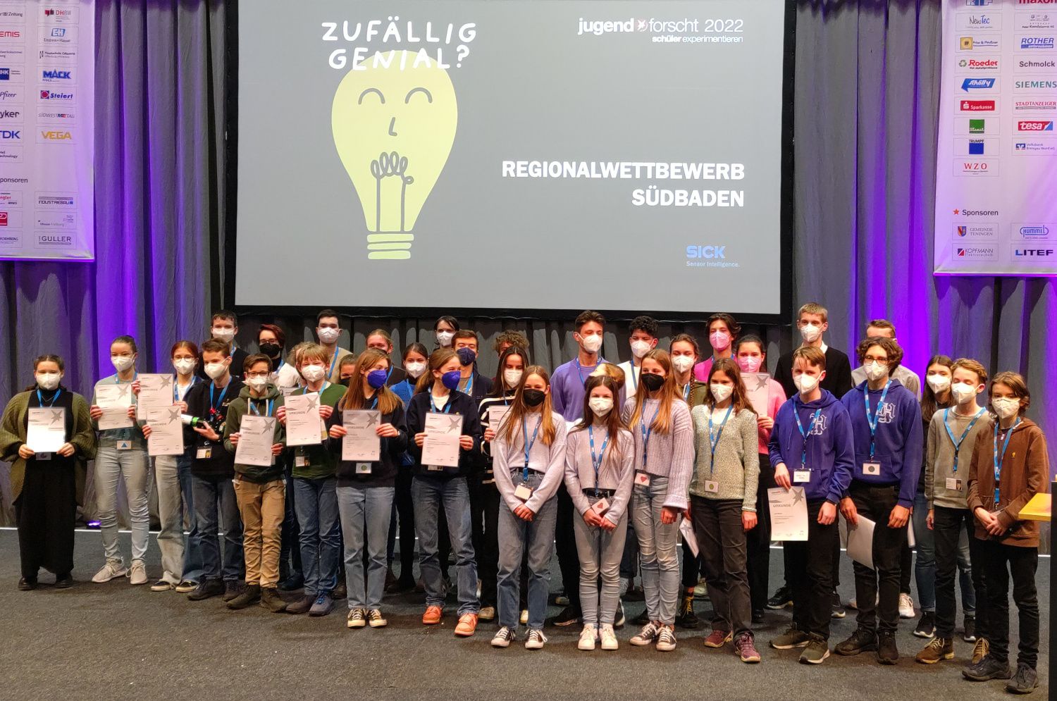 Die Teilnehmenden des Regionalwettbewerbs Südbaden 2022 stehen mit Maske und Urkunde auf der Bühne, auf der Leinwand das Motto „Zufällig genial?“