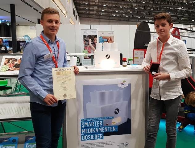 Zwei Jugendliche stehen mit Urkunde und Medaille an ihrem Messestand, davor das Plakat zum smarten Medikamenten-Dosierer