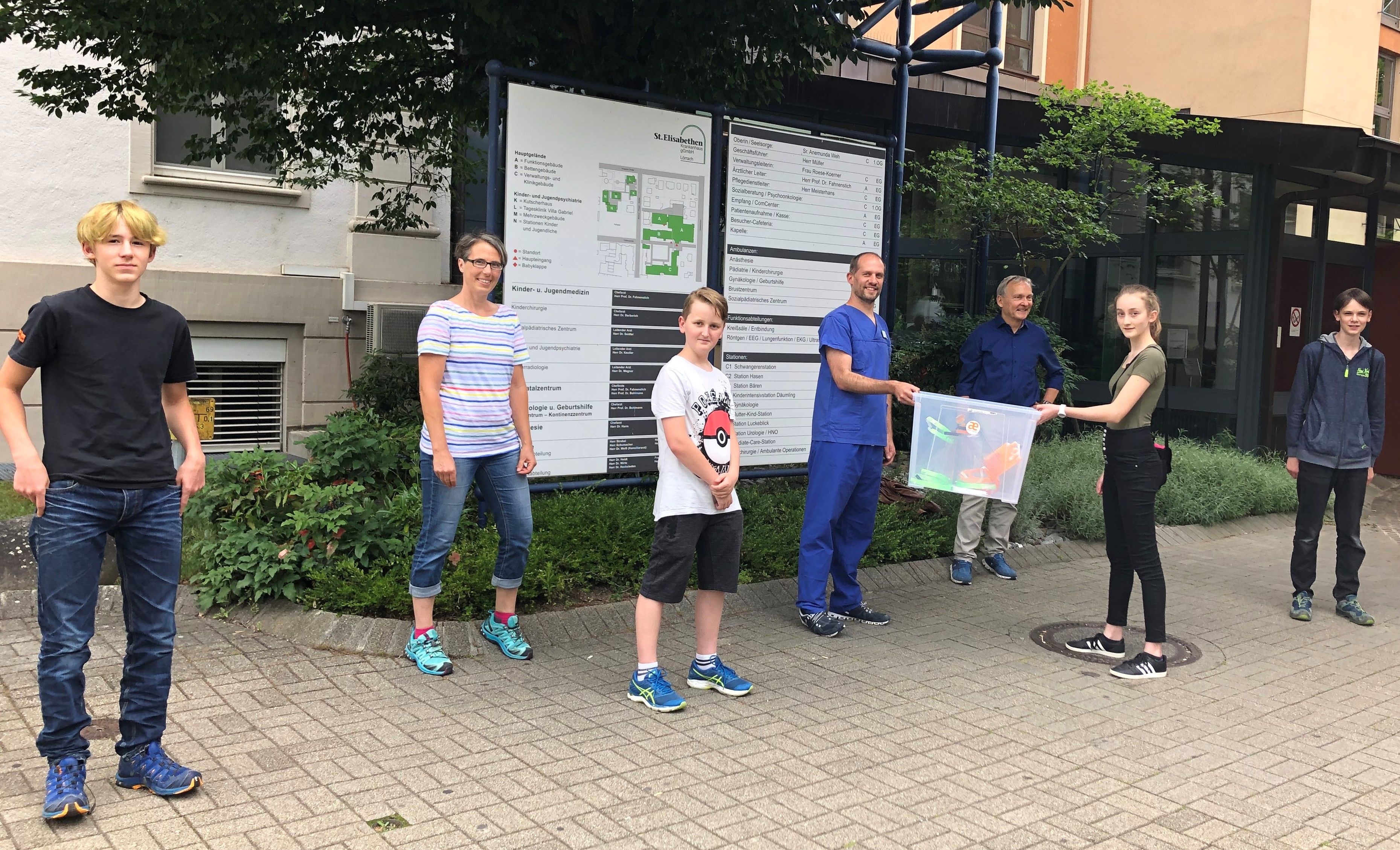 Übergabe einer Kiste mit selbst gedruckten Teilen vor dem Klinikeingang, die Beteiligten stehen mit Abstand nebeneinander