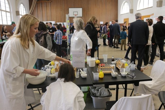 Eine Betreuerin im Laborkittel rührt an einem Mitmachtisch in der vollen Halle eine Mischung an, ein Kind schaut ihr zu