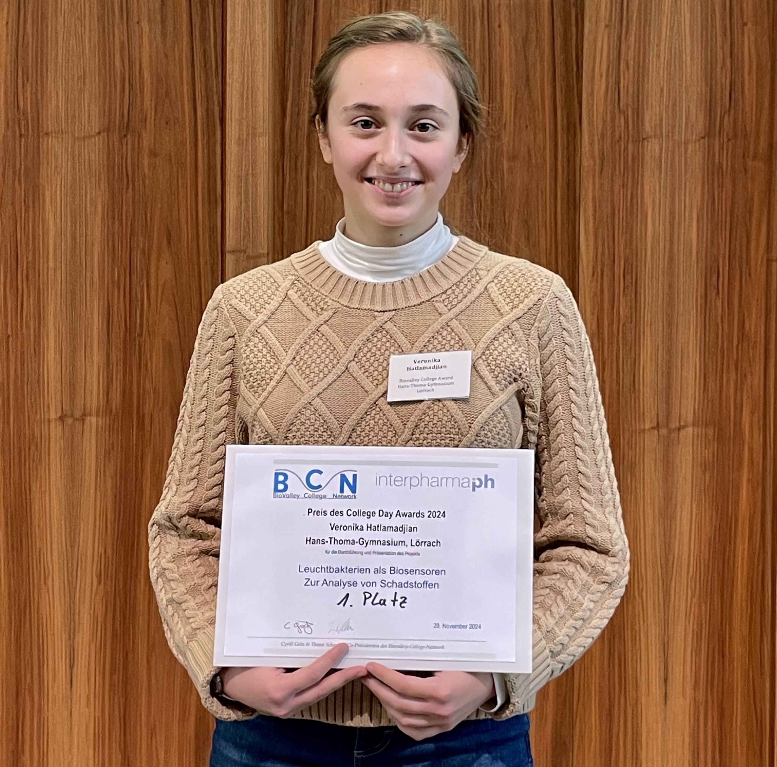 Eine junge Frau hält die Urkunde für den ersten Platz beim College Day Award 2024 vor einer Holzwand