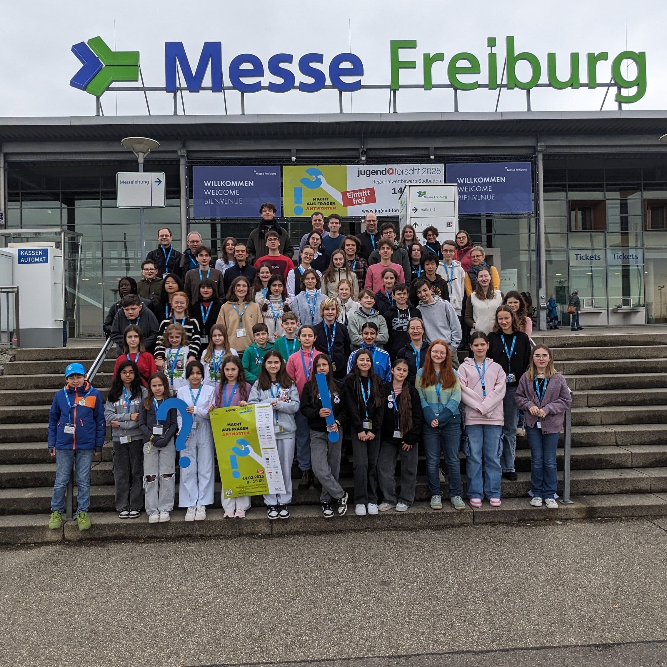 Große Gruppe Kinder, Jugendlicher und Betreuender auf der Treppe der Messe Freiburg vor dem Banner des Jugend-forscht-Regionalwettbewerbs 2025, einige halten blaue Fragezeichen aus Pappe