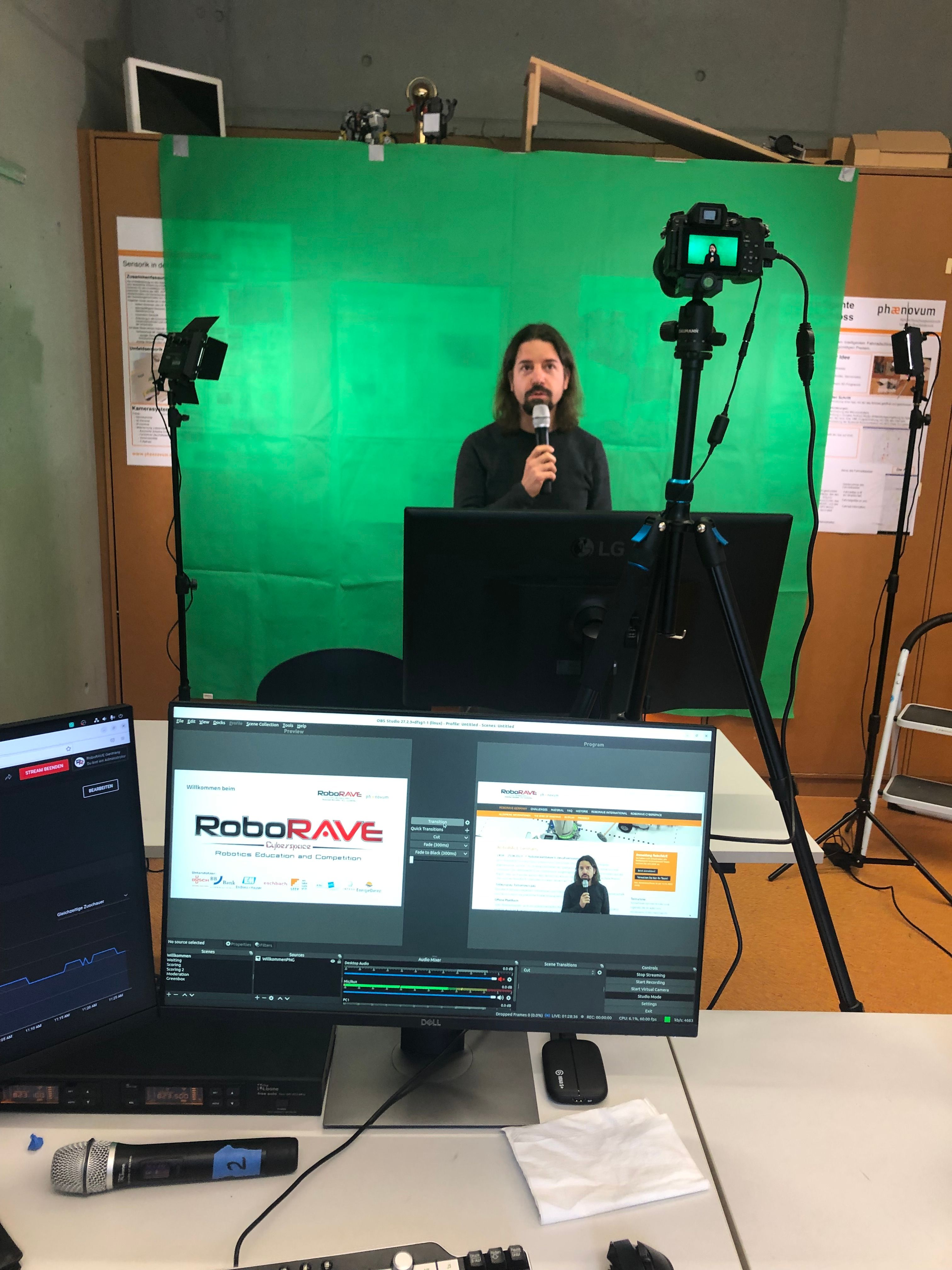 Im improvisierten Studio moderiert ein Mann mit Mikrofon vor der Greenscreen-Wand, auf den Monitoren läuft die Streamsoftware mit dem RoboRAVE-Logo