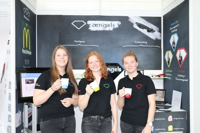 Drei Schülerinnen der Schülerfirma ængels stehen mit bedruckten Tassen vor ihrer Messewand aus Tafelfolie