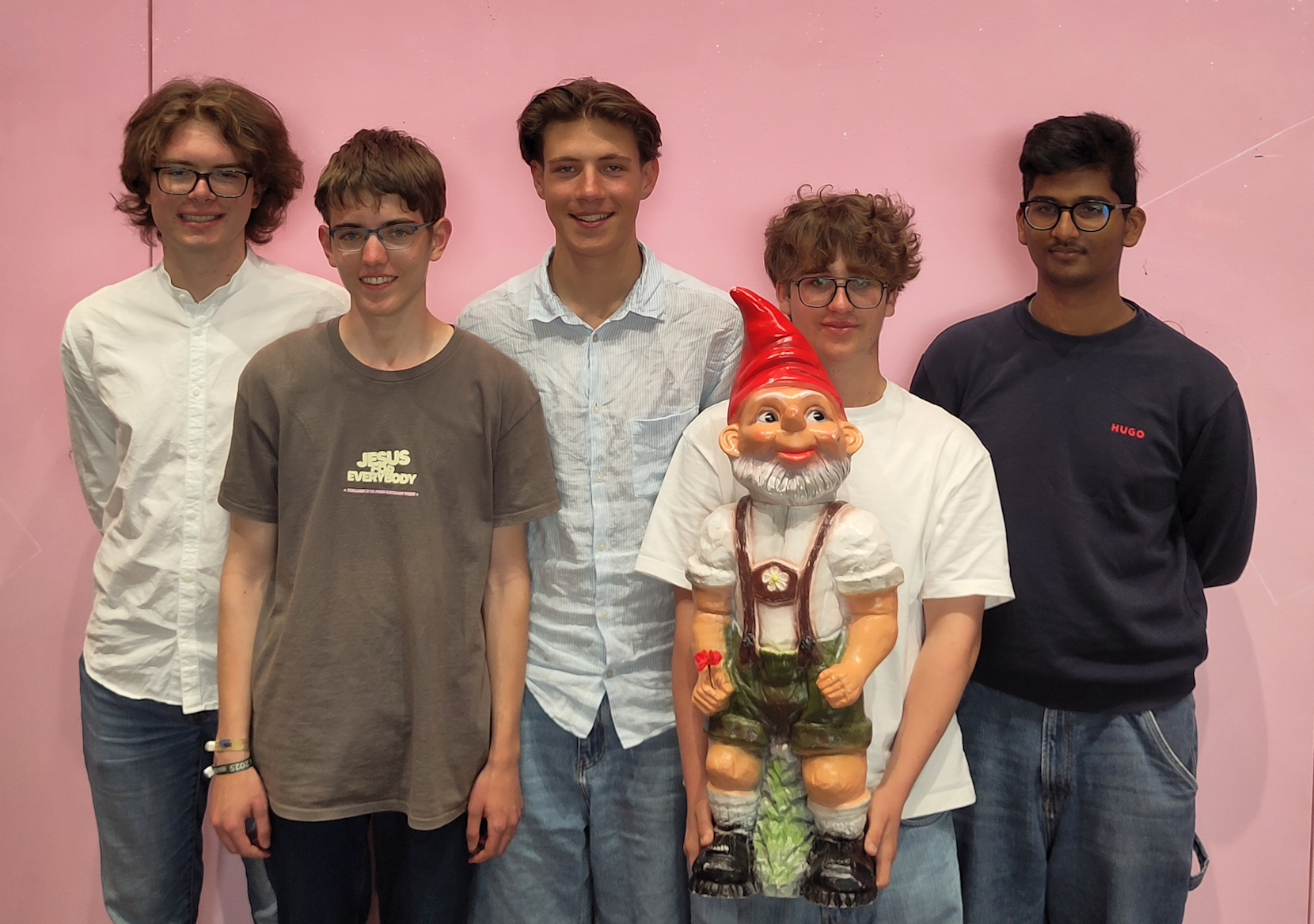 Fünf Jugendliche stehen vor einer rosafarbenen Wand, einer von ihnen hält einen Gartenzwerg in Lederhosen