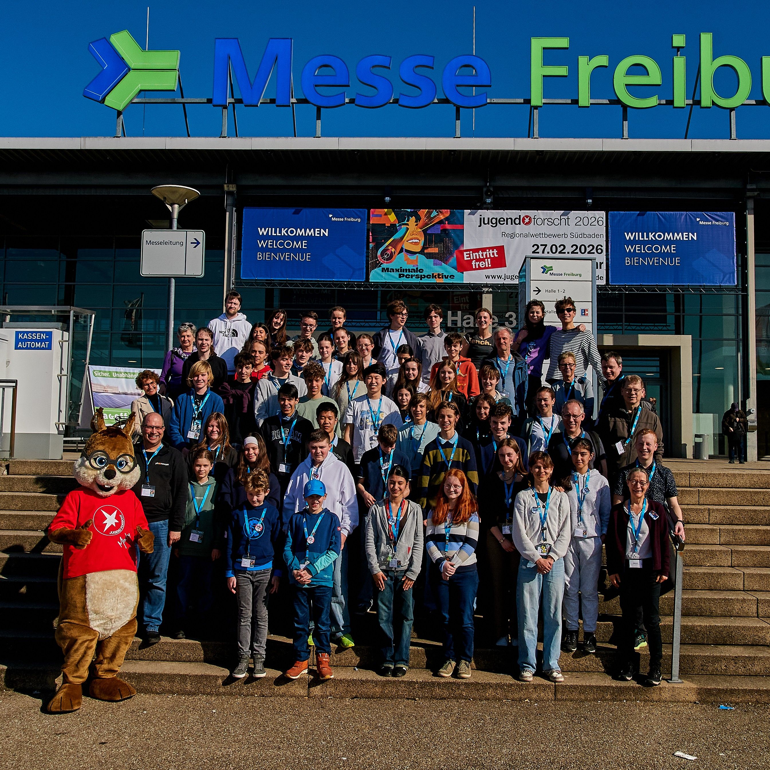 Große Gruppe Jugendlicher und Betreuender auf der Treppe der Messe Freiburg, am Rand ein Eichhörnchen-Maskottchen im roten Shirt