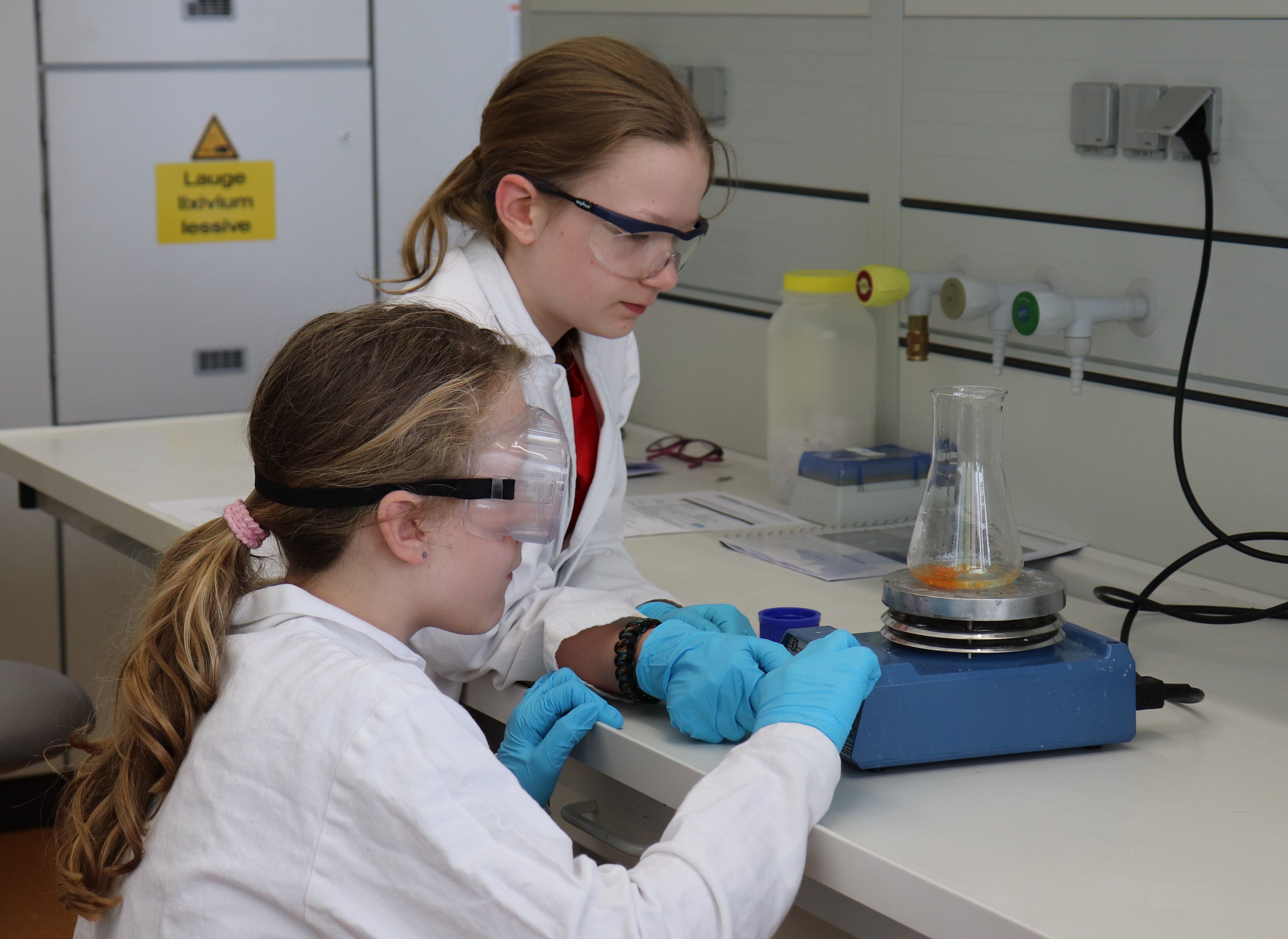 Zwei Mädchen in Laborkittel, Schutzbrille und Handschuhen arbeiten am Labortisch, auf dem Magnetrührer steht ein Kolben mit oranger Lösung