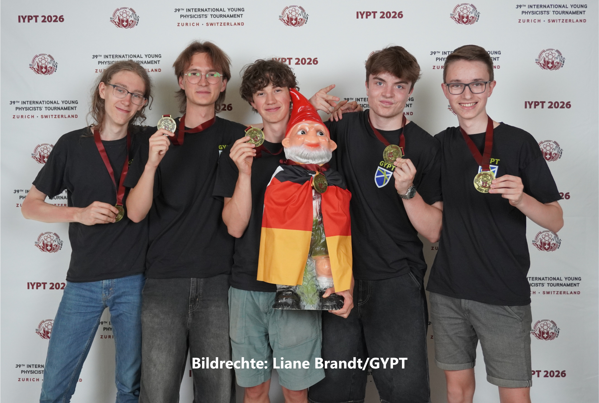 Fünf Jugendliche in schwarzen Shirts halten ihre Goldmedaillen vor der Fotowand des 39. International Young Physicists’ Tournament in Zürich hoch, in ihrer Mitte ein Gartenzwerg mit Deutschlandflagge