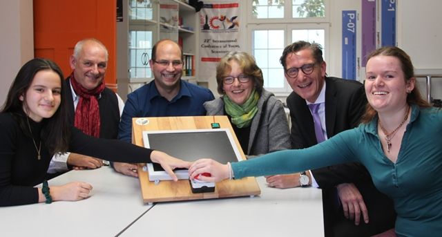 Bei einem Pressegespräch legen sechs Personen lächelnd gemeinsam die Hand auf ein selbst gebautes Gerät mit Holzrahmen und Bildschirm