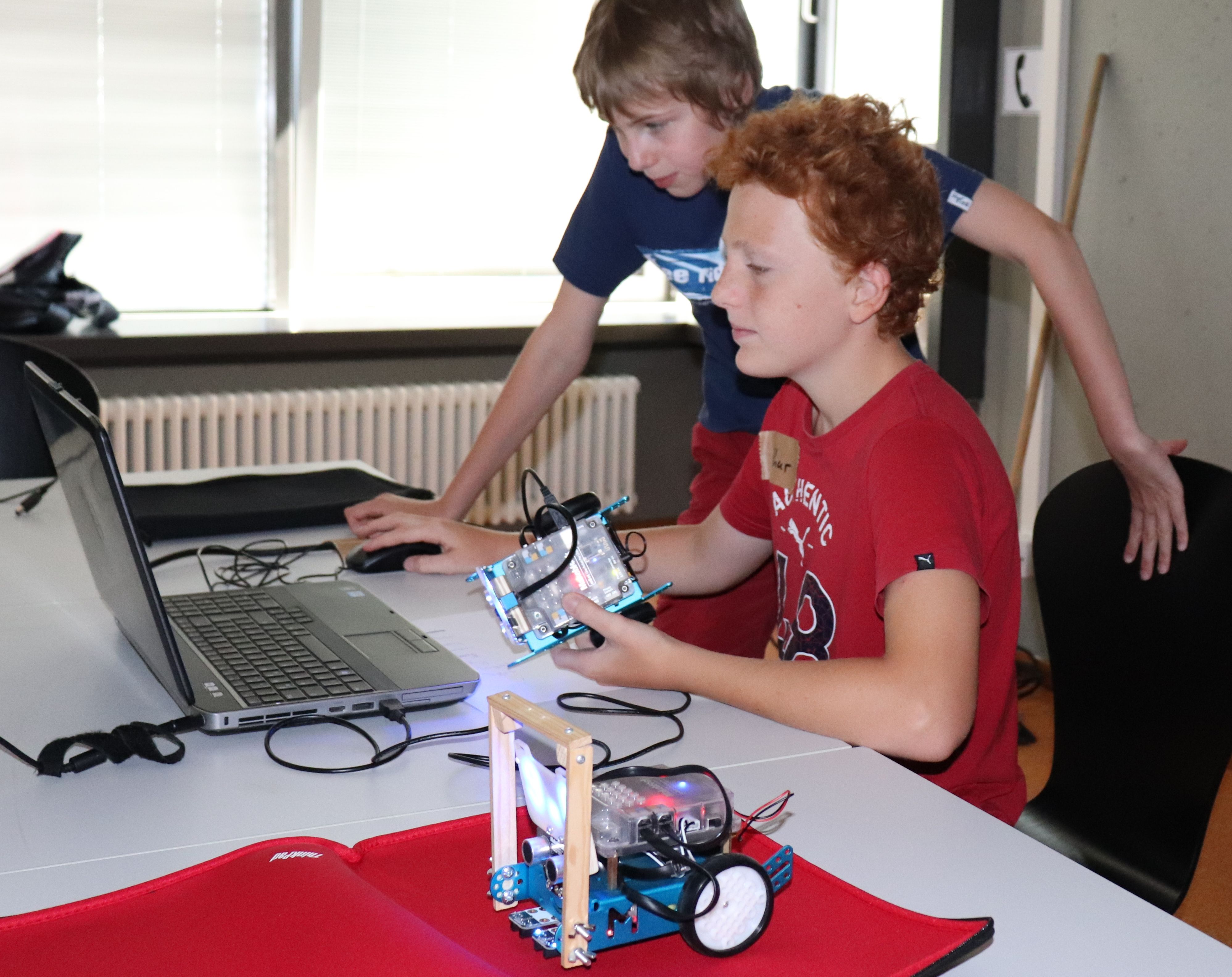 Zwei Jungen im Ferienkurs schauen auf den Laptop, einer hält dabei die Platine seines Roboters in der Hand