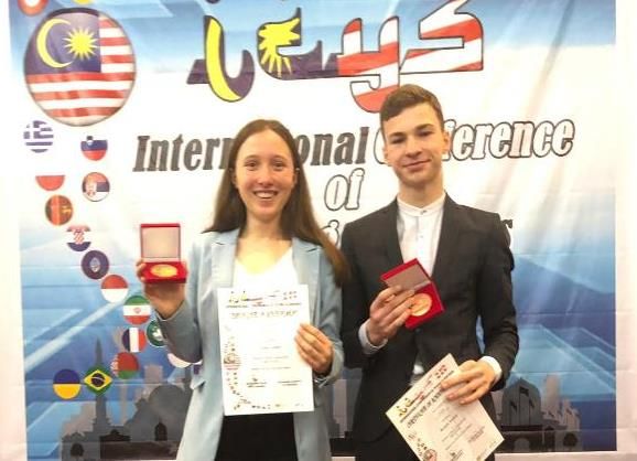 Zwei Jugendliche halten Medaille und Urkunde vor der Flaggenwand der International Conference of Young Scientists