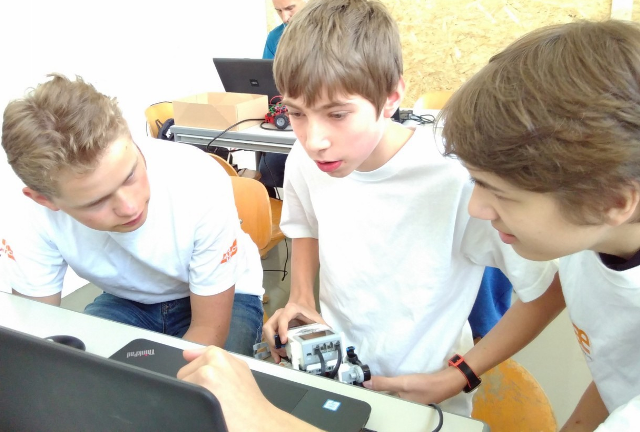 Drei Jungen in weißen T-Shirts beugen sich über einen Laptop, der mittlere hält einen selbst gebauten Roboter in den Händen
