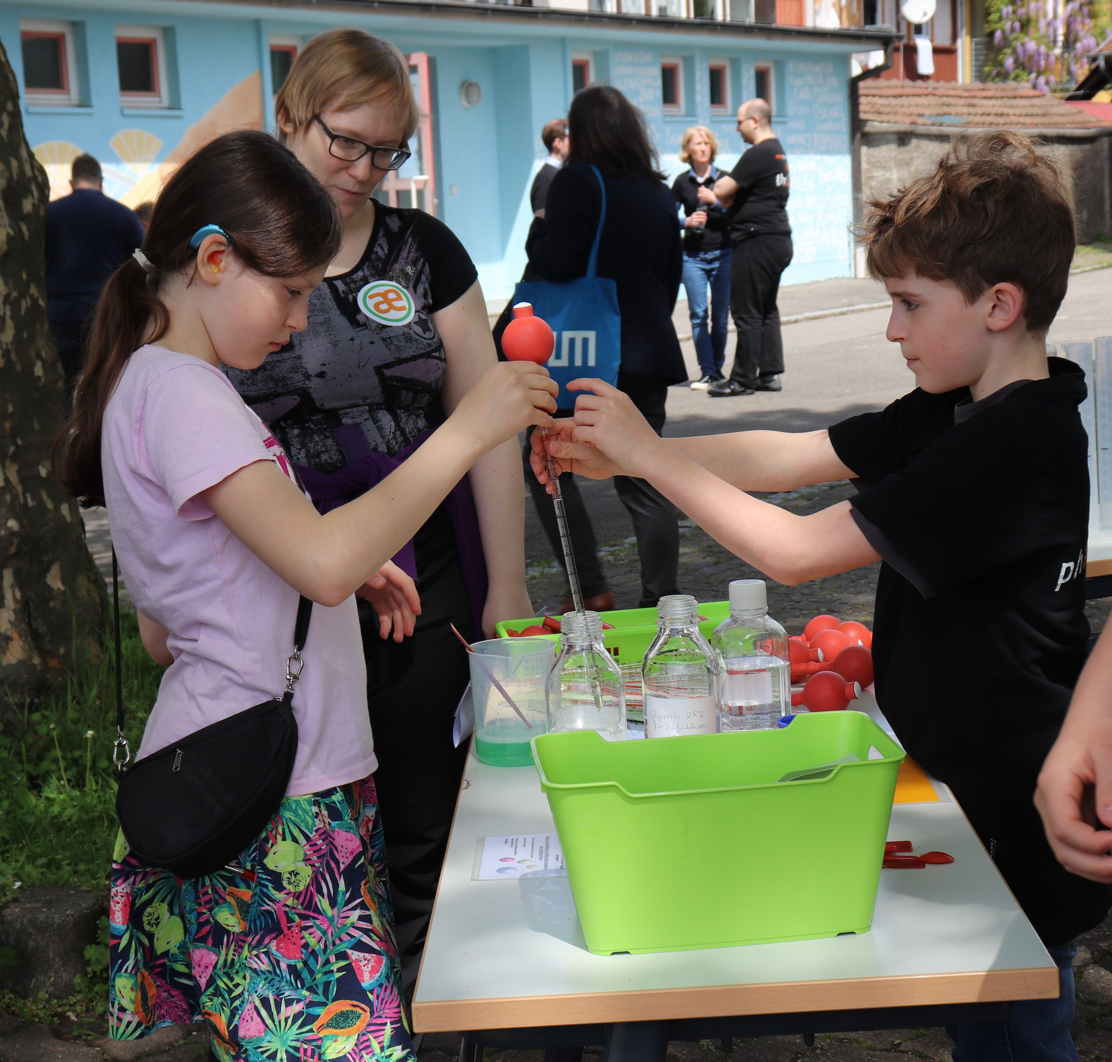 An einem Stand im Freien reicht ein Junge einem Mädchen eine Pipette mit rotem Ball, daneben schaut eine Erwachsene zu