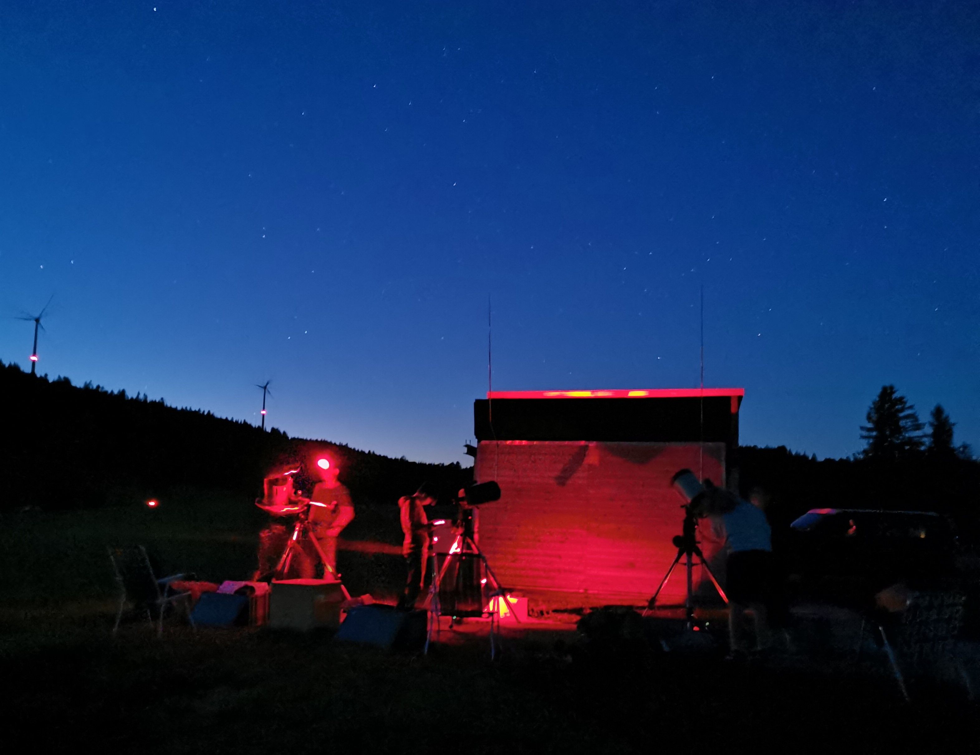 Bei Nacht beobachten mehrere Personen im roten Licht mit Teleskopen neben der Beobachtungshütte, am Horizont stehen Windräder