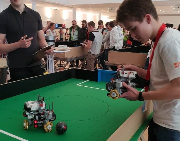 Ein Jugendlicher setzt seinen Lego-Roboter auf das grüne Spielfeld, gegenüber wartet der zweite Roboter beim Roboterfußball
