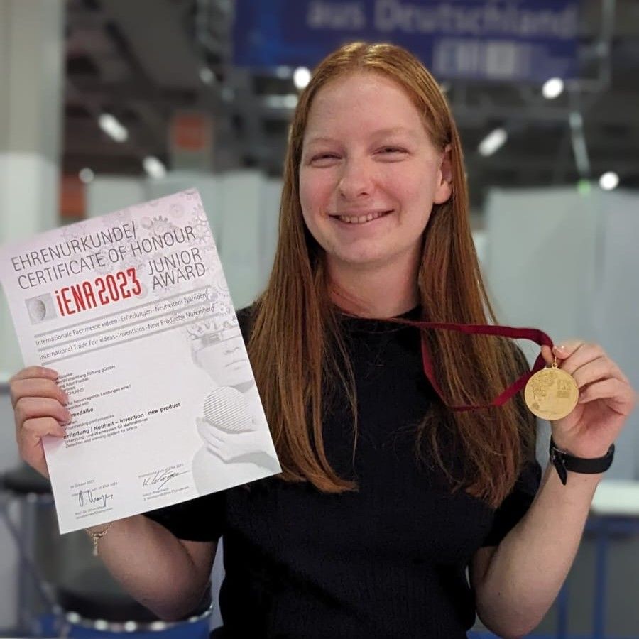Eine junge Frau hält eine Ehrenurkunde der Erfindermesse iENA 2023 und eine goldene Medaille in die Kamera