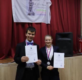 Zwei Jugendliche halten unter dem Banner des ICYS 2018 in Belgrad ihre Urkunden in die Kamera, eine von ihnen trägt eine Medaille um den Hals