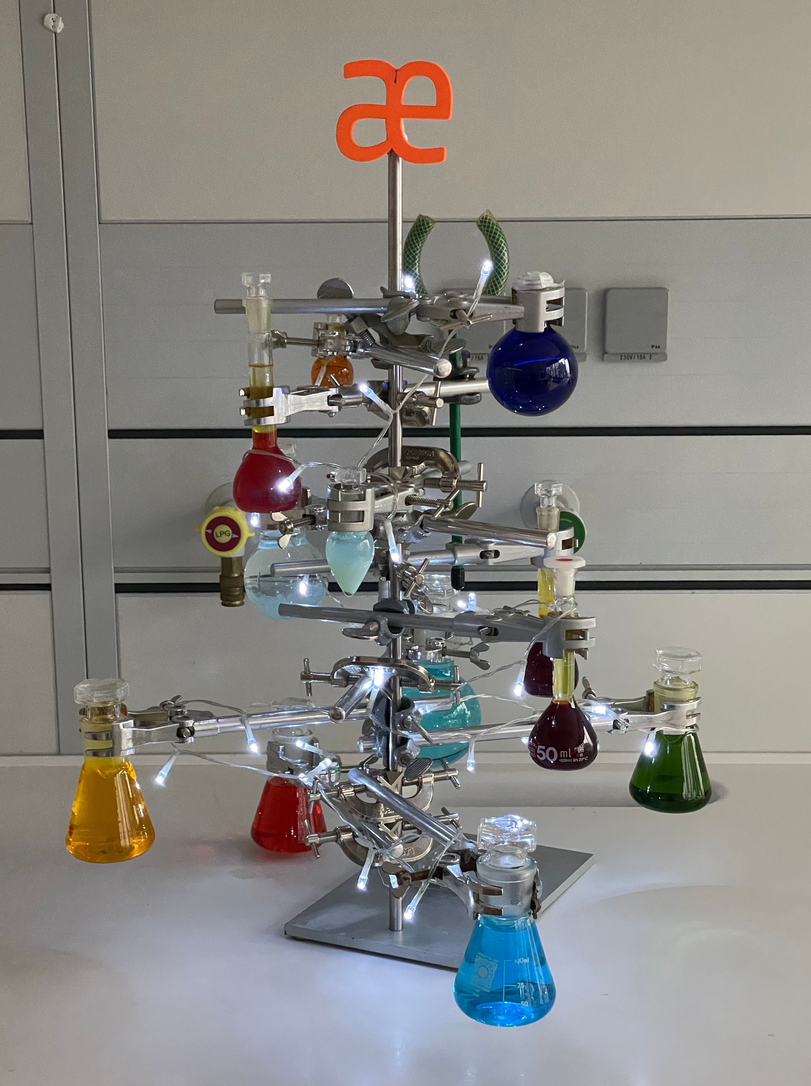 Weihnachtsbaum aus Laborstativen und Klemmen, behängt mit Kolben voller bunter Flüssigkeiten und einer Lichterkette, obenauf das orange æ