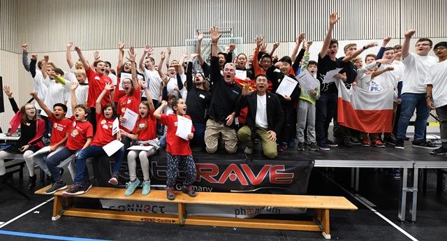 Eine große Gruppe Kinder und Betreuender jubelt mit Urkunden hinter einem RoboRAVE-Banner in einer Sporthalle