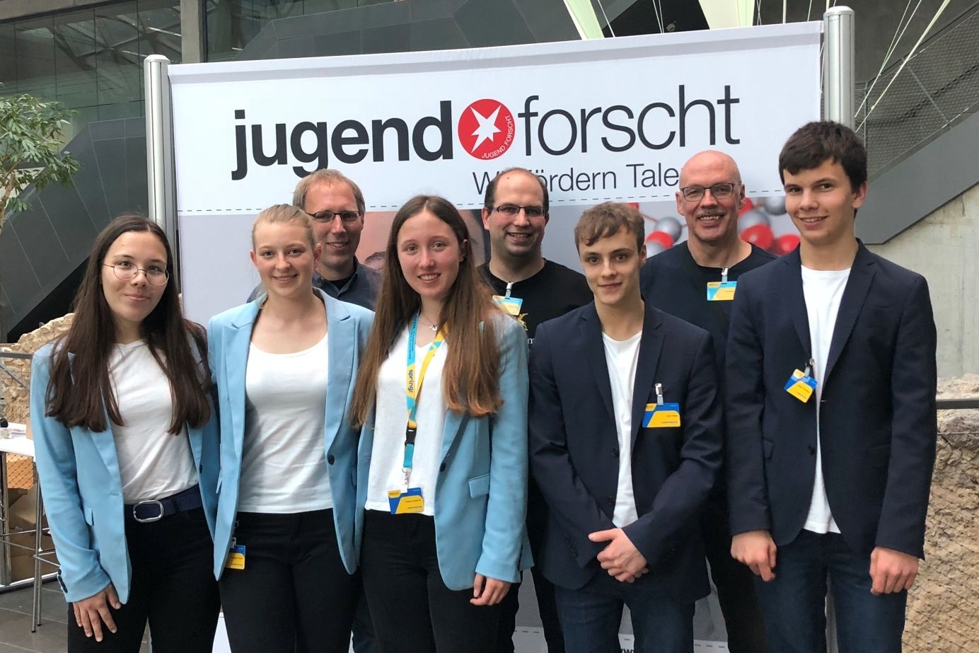 Jungforscherinnen und Jungforscher in hellblauen Sakkos stehen mit ihren Betreuern vor der Wand des Wettbewerbs Jugend forscht