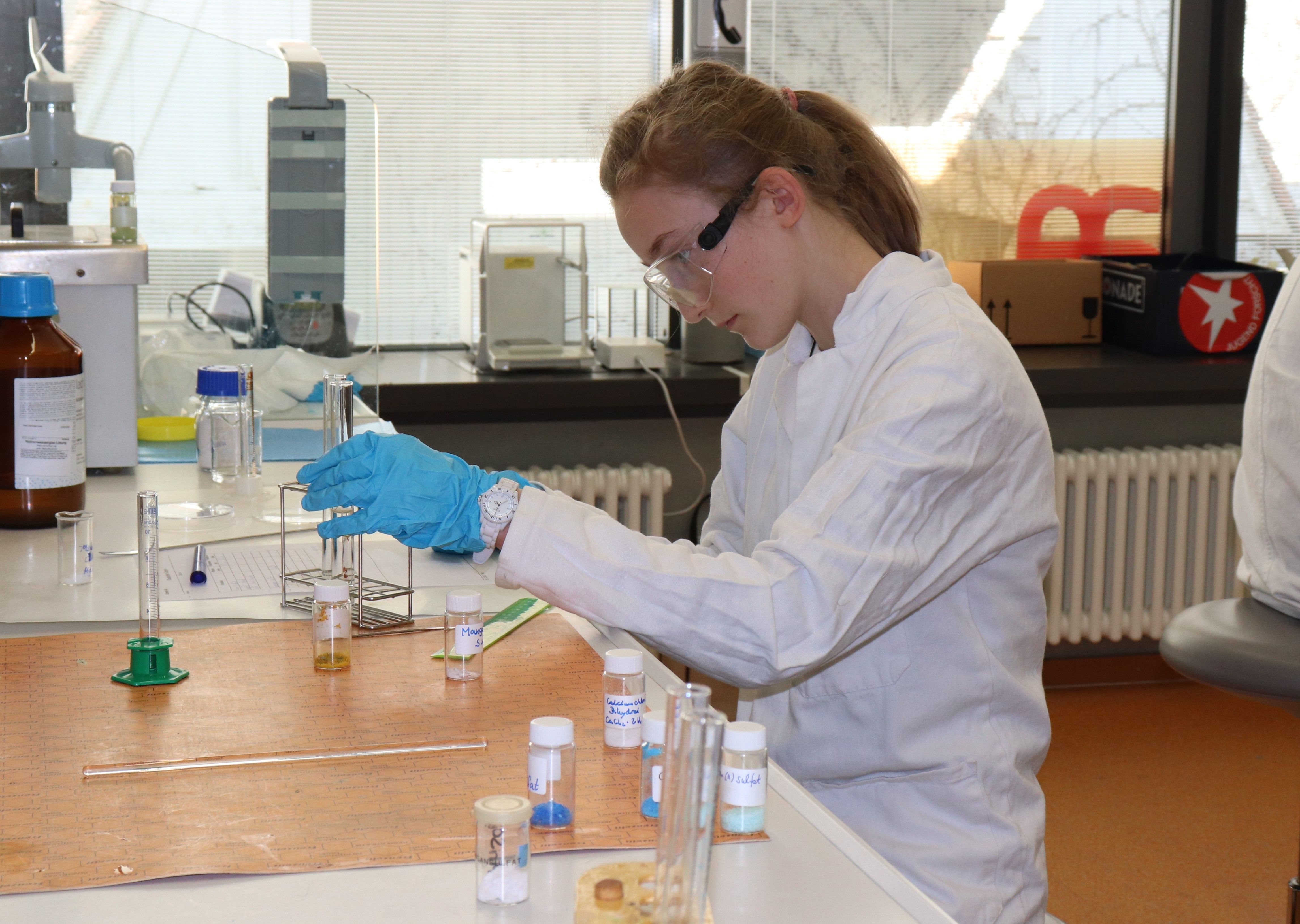 Eine Schülerin in Laborkittel, Schutzbrille und Handschuhen setzt am Labortisch Proben in Reagenzgläsern an