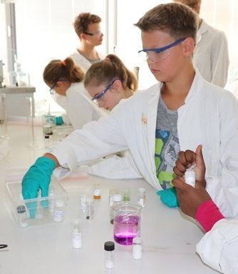 Kinder in Laborkitteln und Schutzbrillen pipettieren am Labortisch, im Vordergrund ein Becherglas mit pinker Lösung