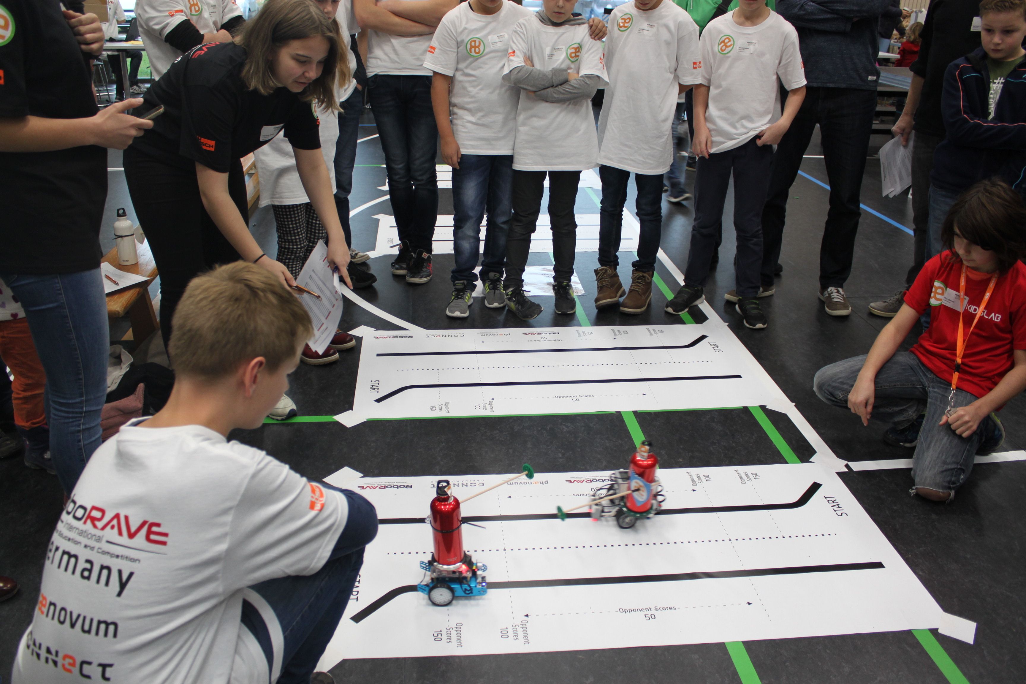 Zwei selbst gebaute Roboter treten auf einer Papierbahn am Hallenboden gegeneinander an, Teams und Publikum schauen zu