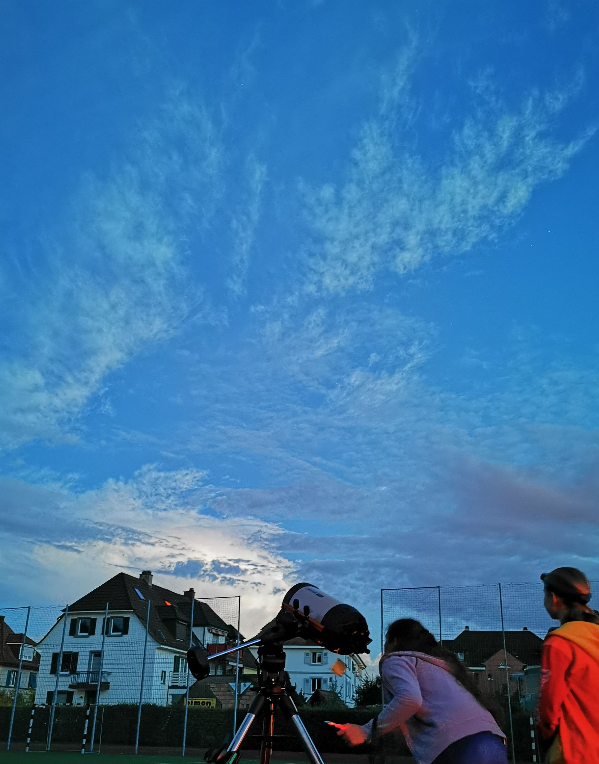 Auf einem Sportplatz steht in der Abenddämmerung ein Teleskop, zwei Jugendliche richten es aus