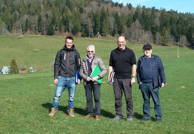 Vier Männer stehen mit Unterlagen unter dem Arm bei einem Ortstermin auf einer Wiese vor dem Wald