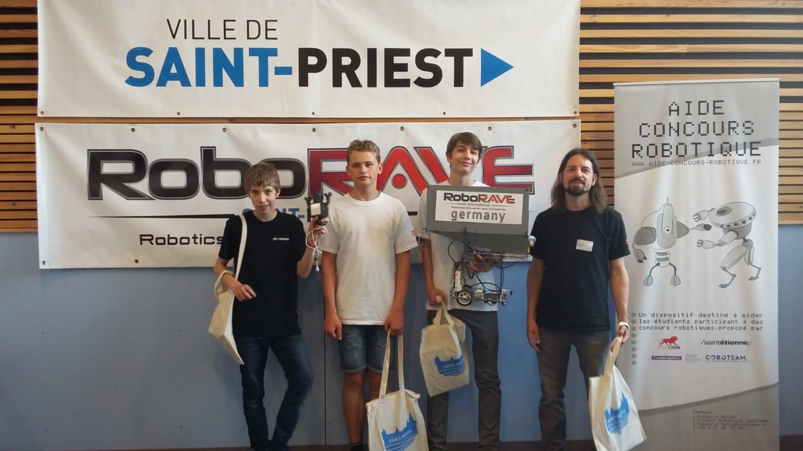 Vier Teilnehmer stehen mit ihrem selbst gebauten Roboter und Stoffbeuteln vor den Bannern des RoboRAVE in Saint-Priest