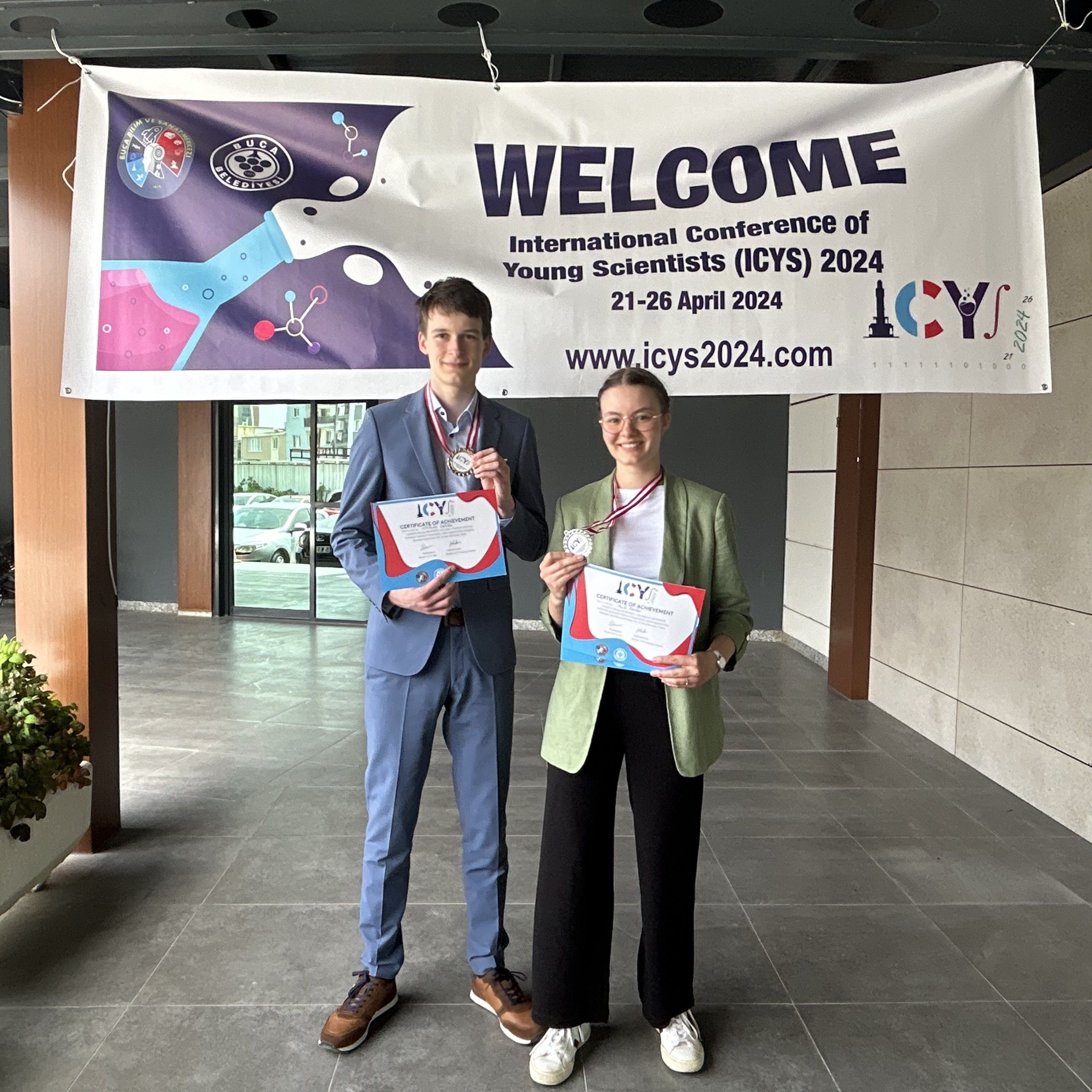 Zwei Jugendliche mit Medaillen und Urkunden unter dem Willkommensbanner der International Conference of Young Scientists 2024