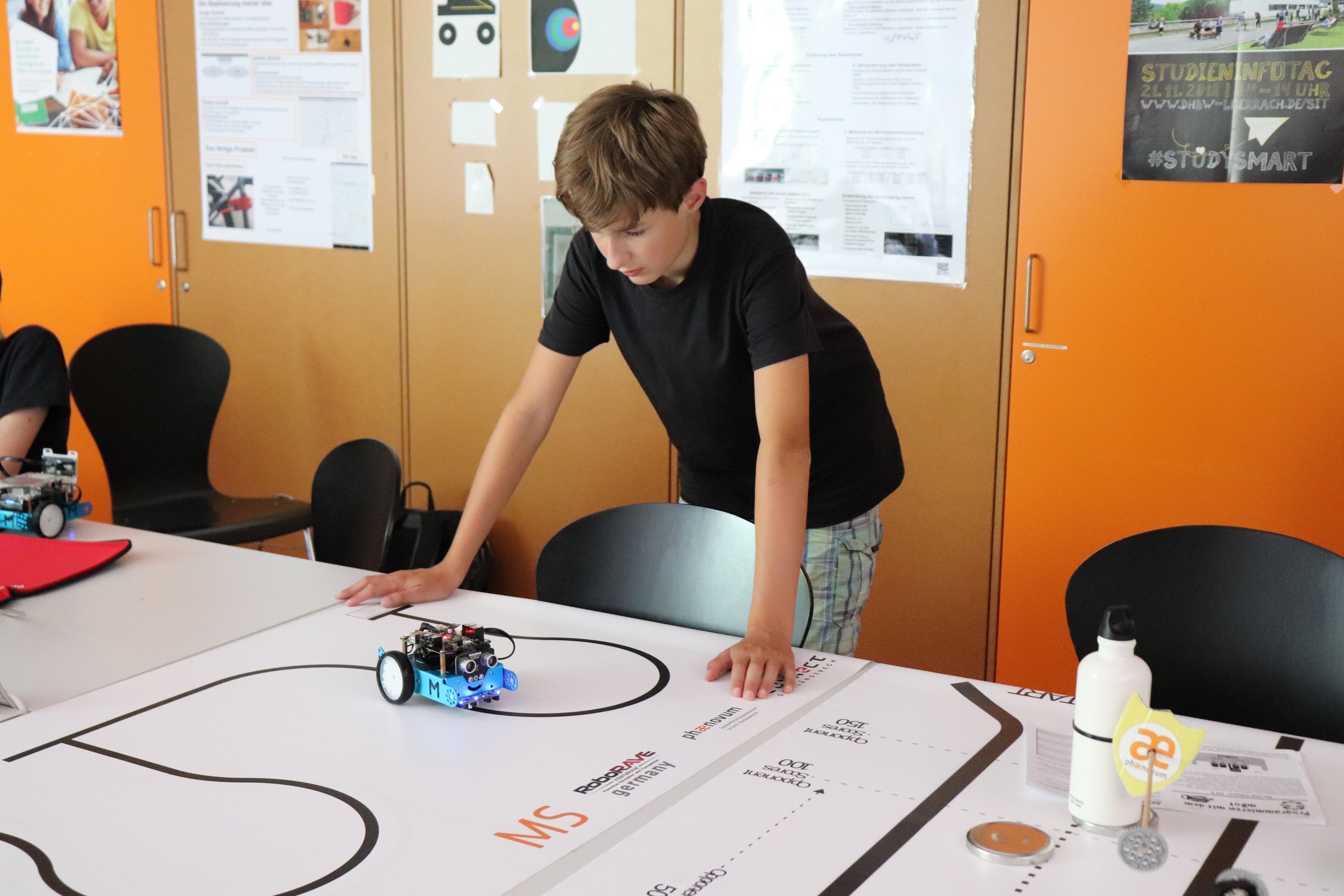 Ein Junge beugt sich über eine weiße Wettbewerbsbahn mit schwarzer Linie, auf der ein kleiner blauer Roboter steht
