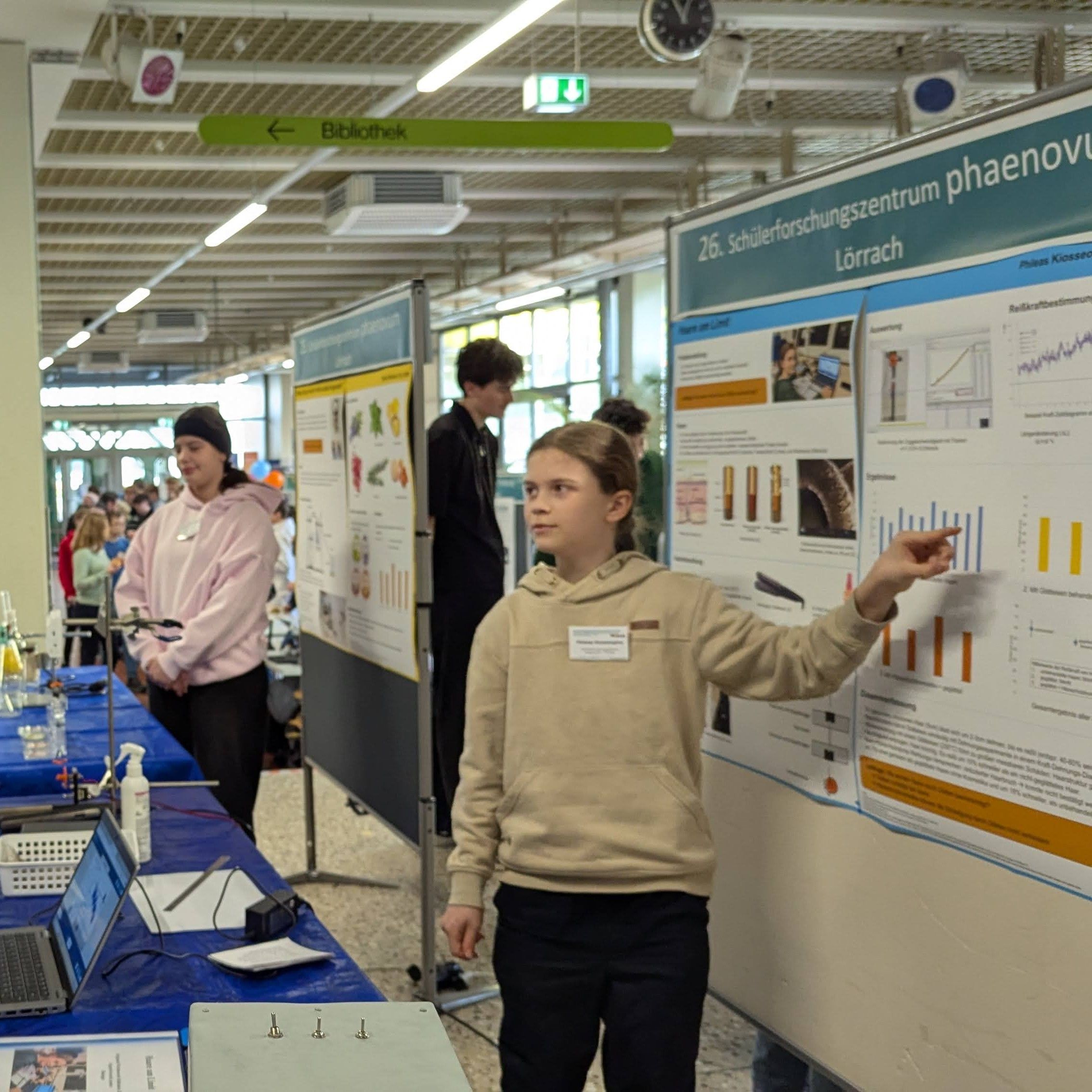 Eine Schülerin zeigt bei einer Posterausstellung auf ein Diagramm ihres Projektplakats, im Hintergrund weitere Stände