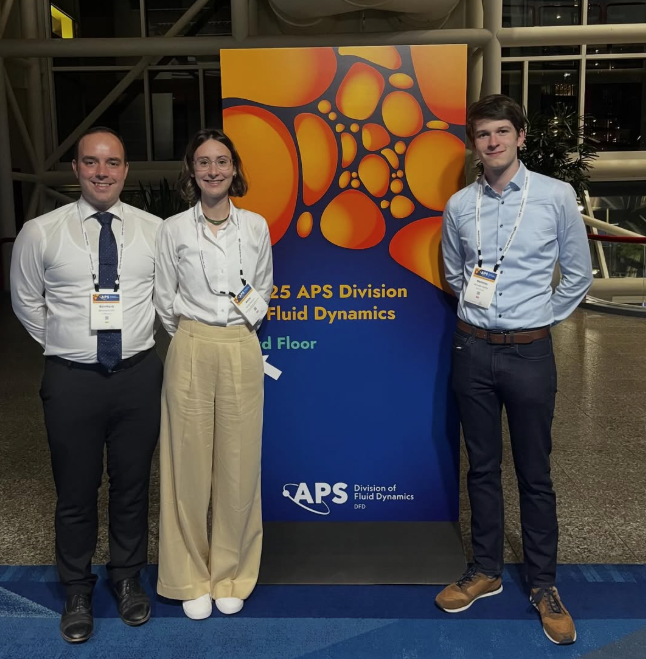 Drei junge Leute mit Konferenzausweisen stehen vor dem Aufsteller der APS Division of Fluid Dynamics 2025