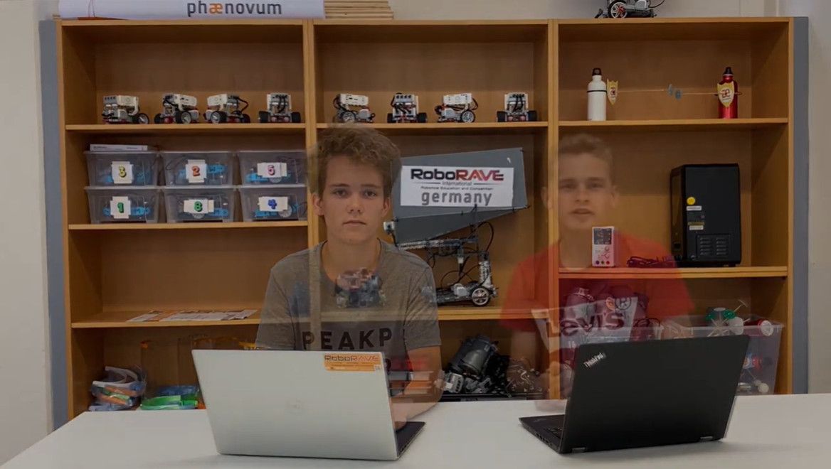 Standbild aus dem Erklärvideo: ein Jugendlicher sitzt mit zwei Laptops vor den Regalen voller Roboter, darüber ist eine zweite Aufnahme geblendet