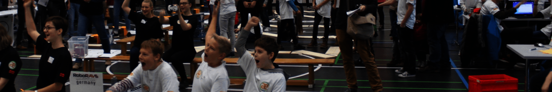 Kinder und Jugendliche jubeln mit erhobenen Armen an den Wettbewerbsbahnen des RoboRAVE in einer Sporthalle