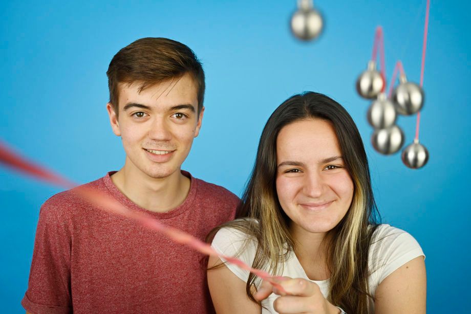 Ein Jugendlicher und eine Jugendliche vor blauem Hintergrund, daneben ein Pendel aus Metallkugeln an roten Fäden, von dem sie einen Faden in die Kamera halten